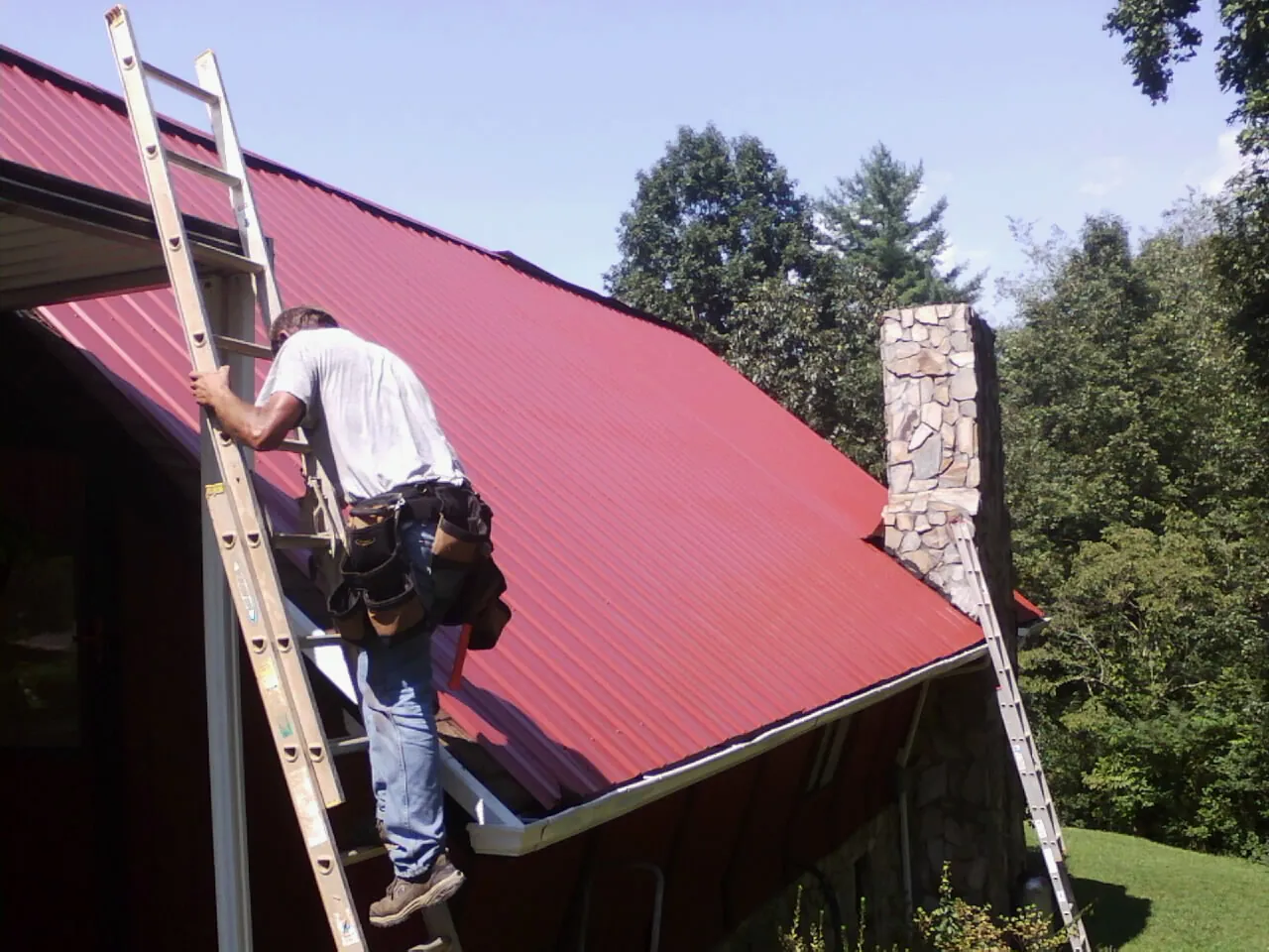 Roofer on a ladder beside a red metal roof and stone chimney — Soffit & Fascia Repair by Roofing Brothers in Woodbridge